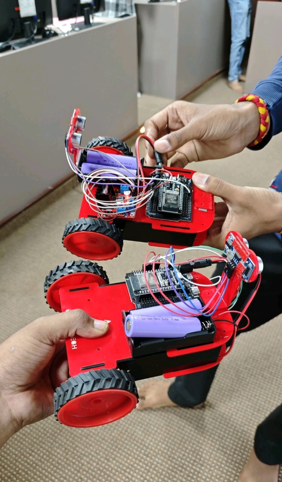 Two red DIY robot chassis held over a lab floor, showing ESP32 boards and ultrasonic sensors