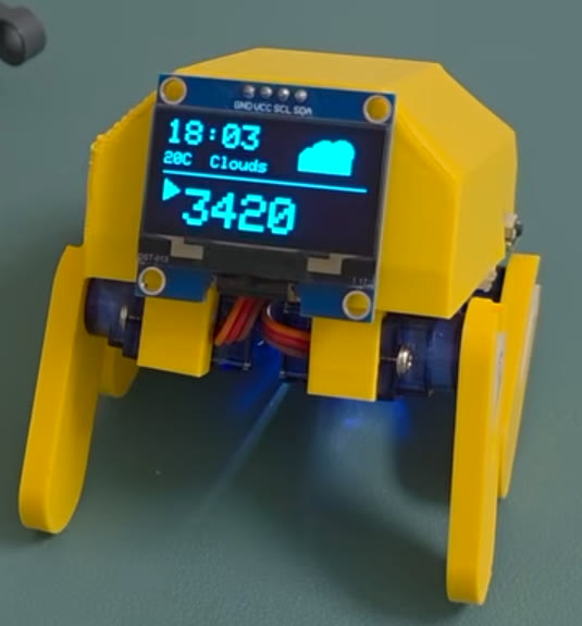 Close-up of a yellow ESP32 walking robot with OLED status screen and blue micro-servos